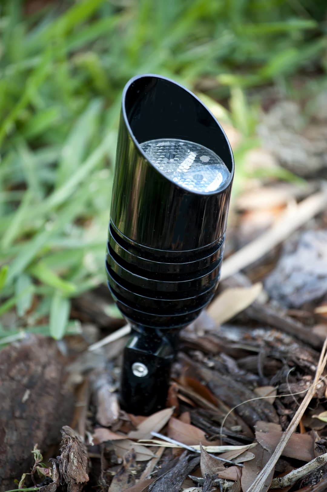 Black outdoor spotlight installed in mulch, surrounded by green grass and leaves.