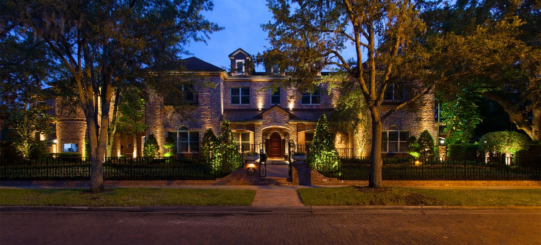 Elegant brick mansion lit up at night, showcasing lush landscaping and a welcoming entrance.