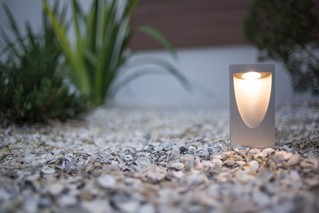 Modern outdoor LED spotlight illuminating gravel garden with green plants in background.