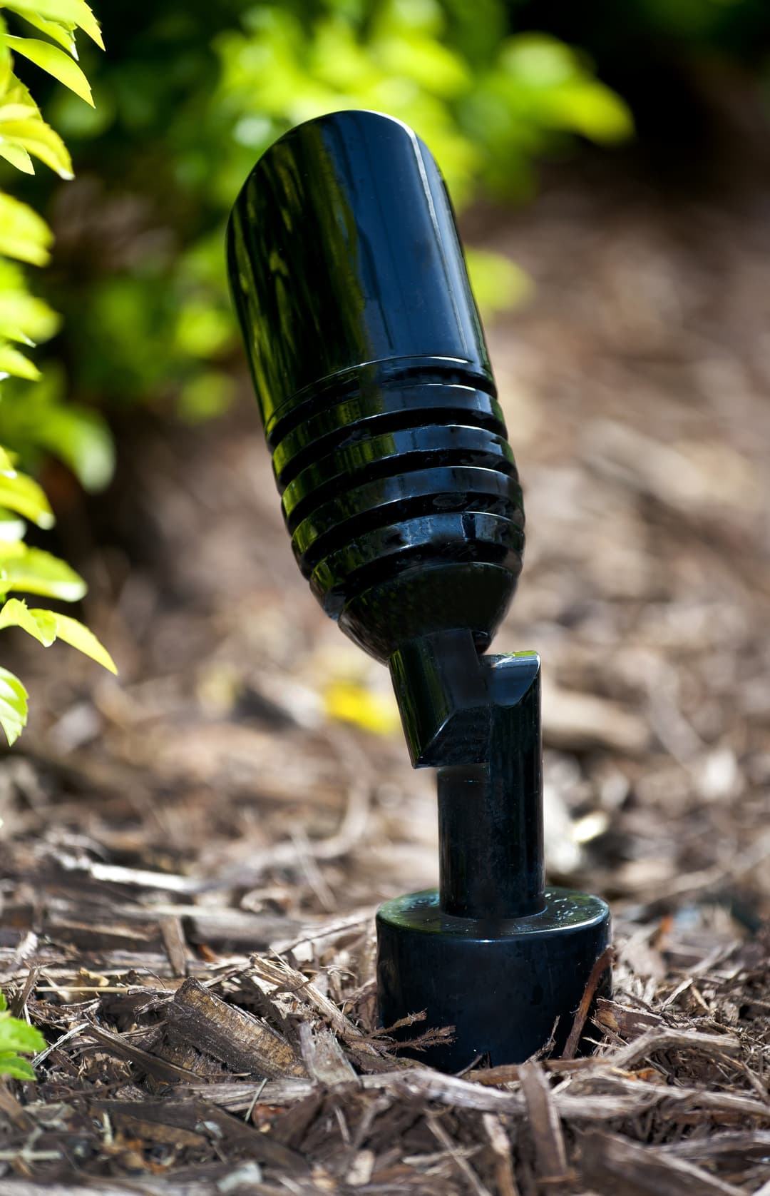 Black landscape light fixture positioned among mulch and plants in a garden setting.