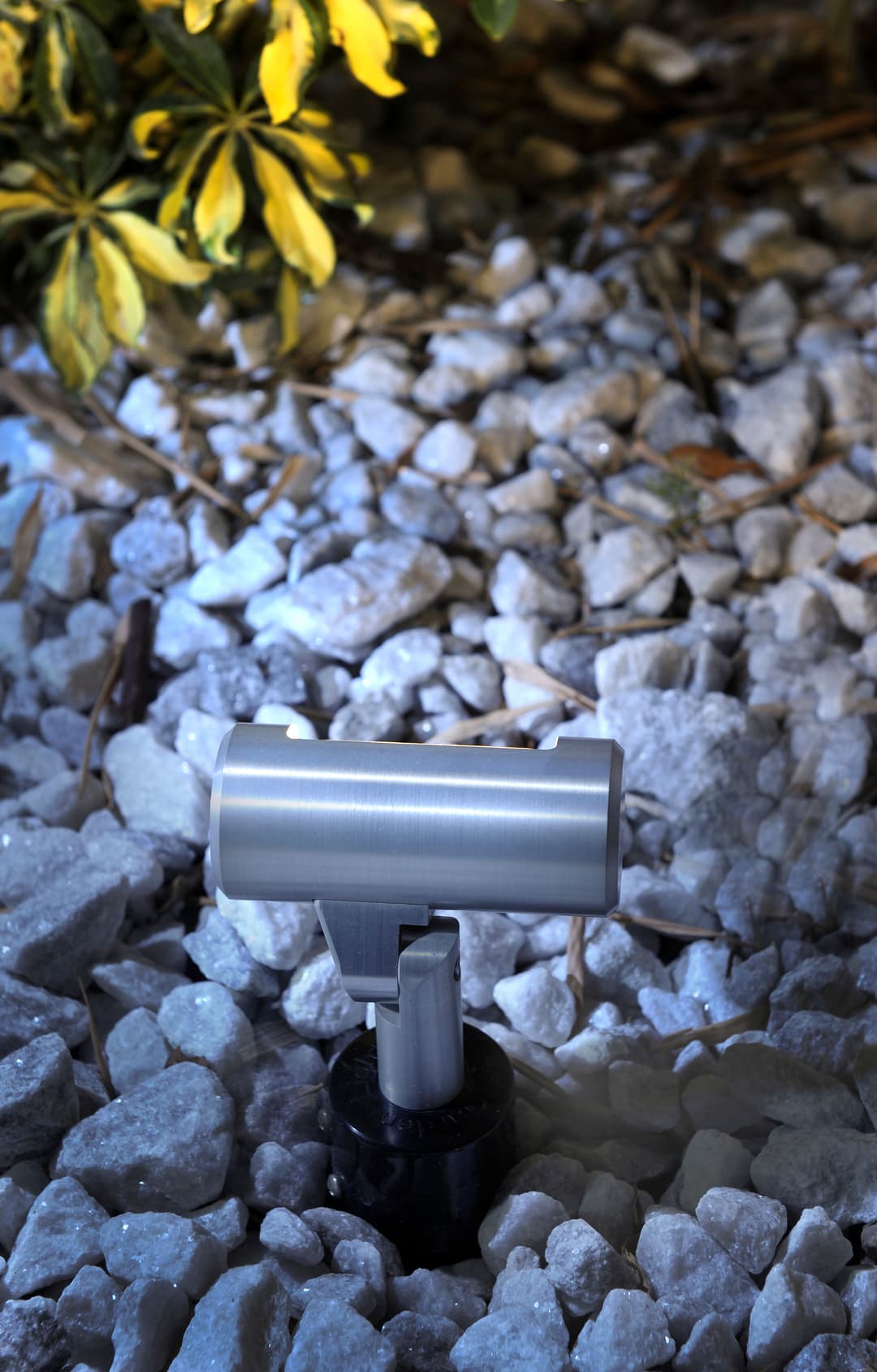 Metal landscape light on gravel, illuminating nearby foliage at night.