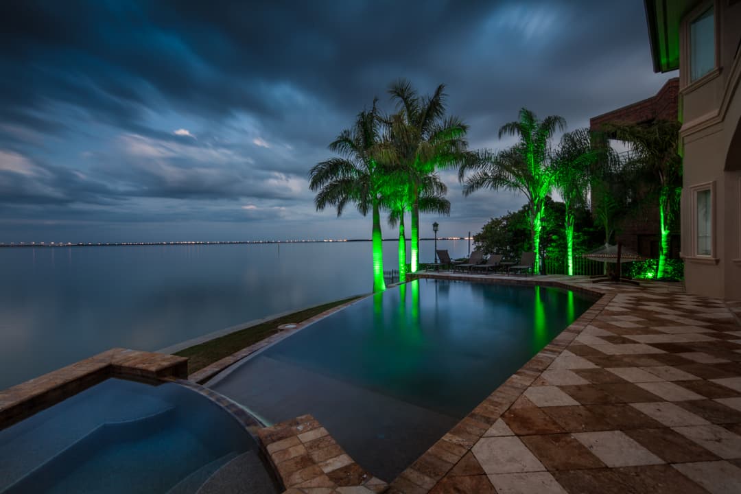 Luxury infinity pool with green illuminated palm trees and serene waterfront view at dusk.
